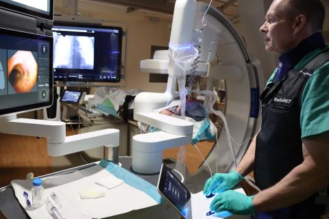 A man in green gloves and a black vest works in a medical room with screens and equipment.