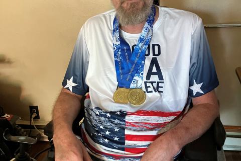 A man with a beard in a wheelchair wearing a USA flag t-shirt and medal.
